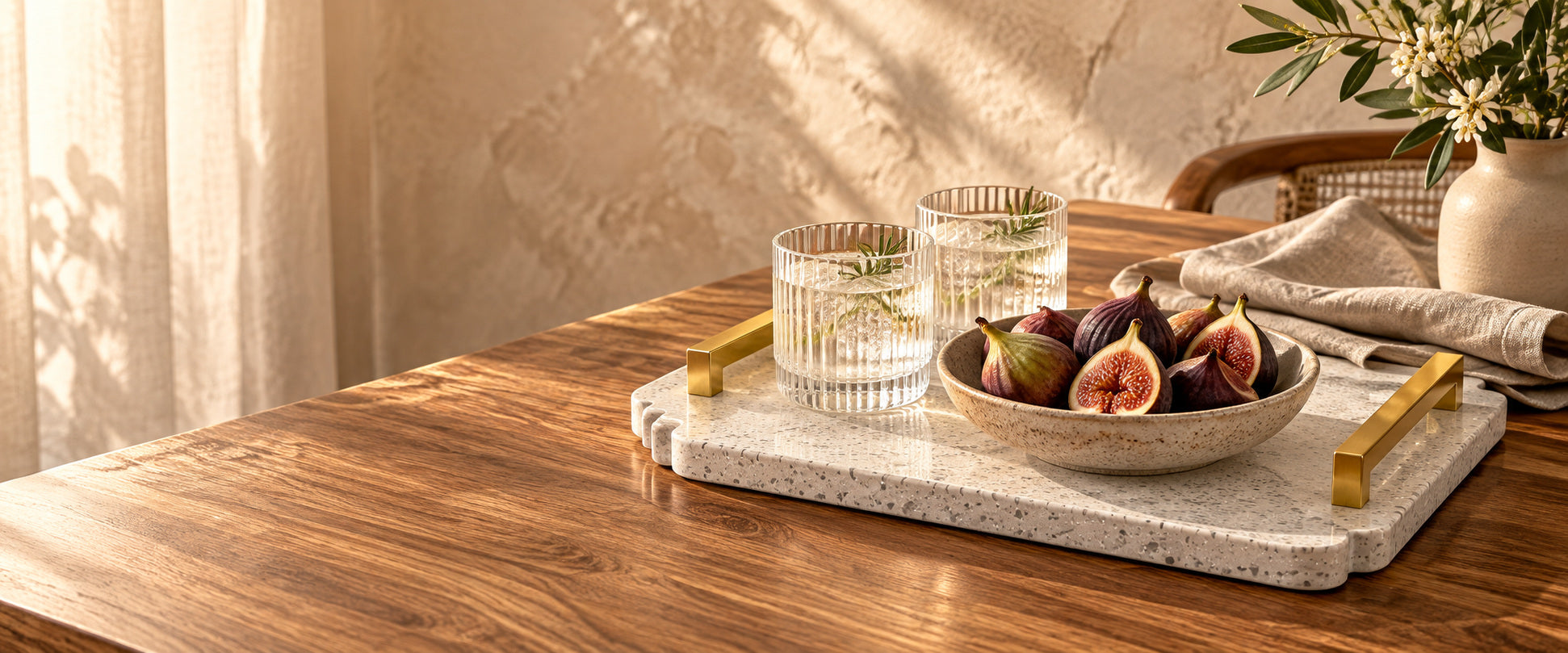 White stone tray with gold handles, glasses, figs and linen on a warm walnut table.